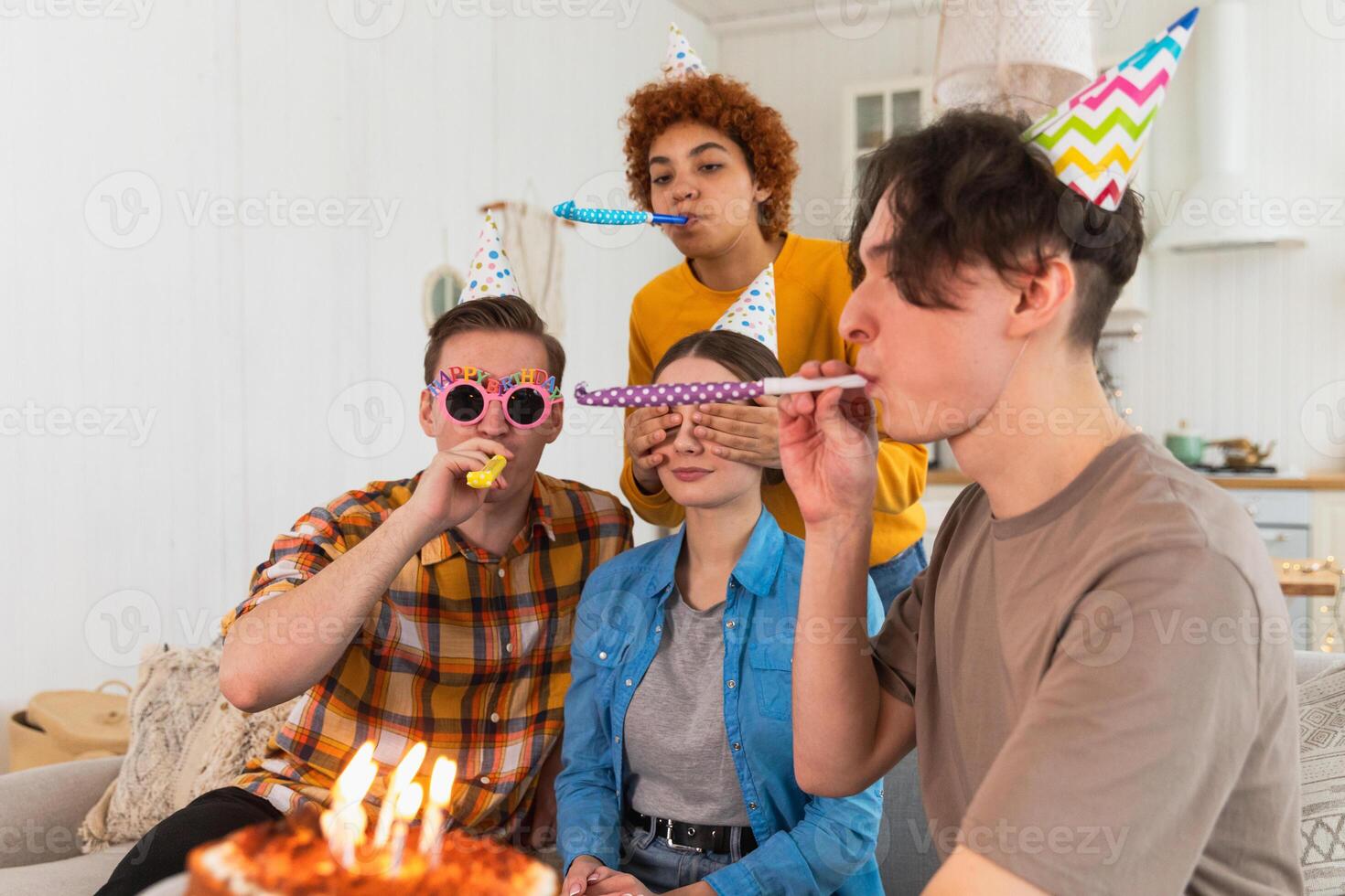 Make a wish. Woman wearing party cap blowing out burning candles on birthday cake. Happy Birthday party. Group of friends wishes girl happy birthday. People celebrating birthday with party at home photo