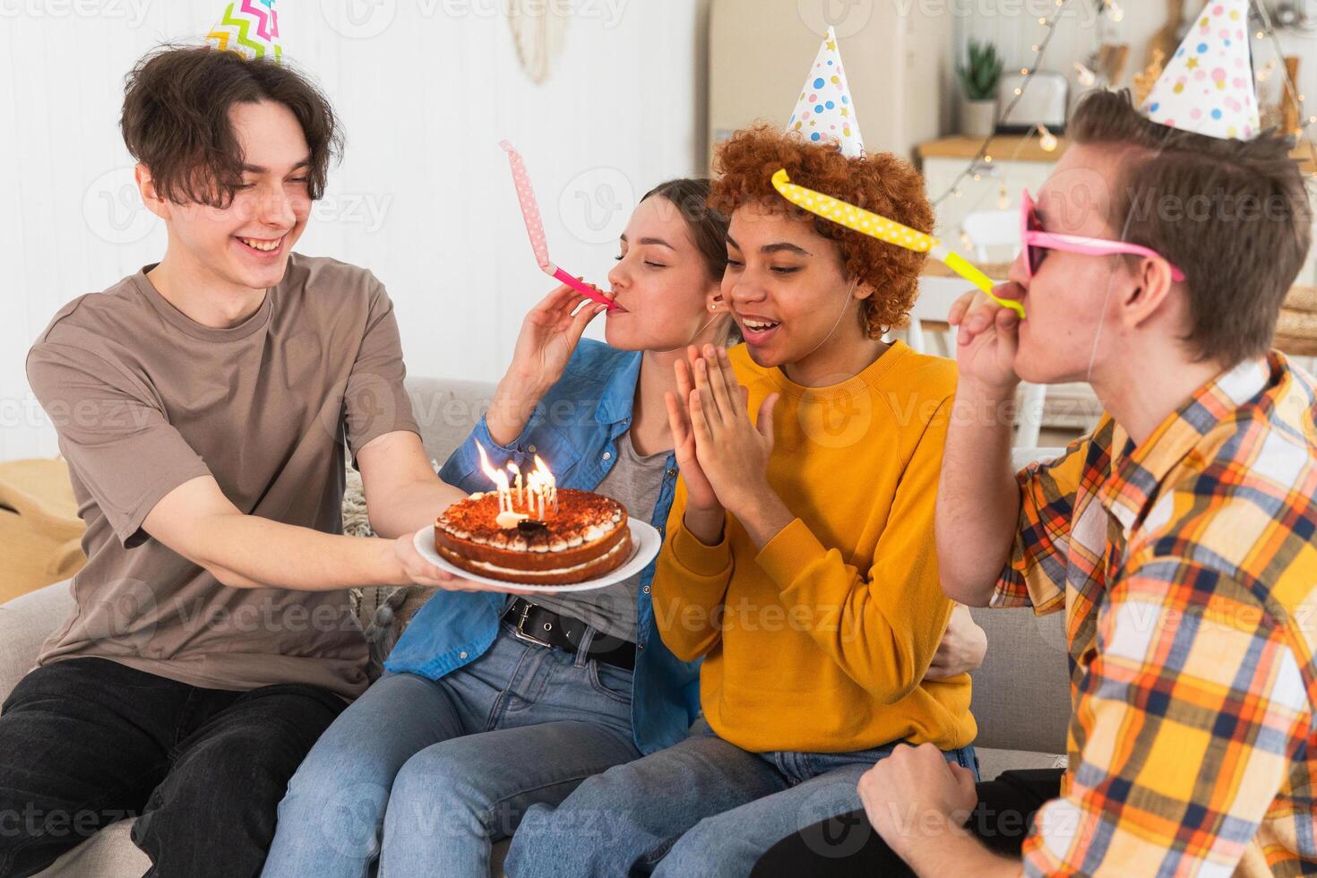 Make a wish. Woman wearing party cap blowing out burning candles on birthday cake. Happy Birthday party. Group of friends wishes girl happy birthday. People celebrating birthday with party at home photo
