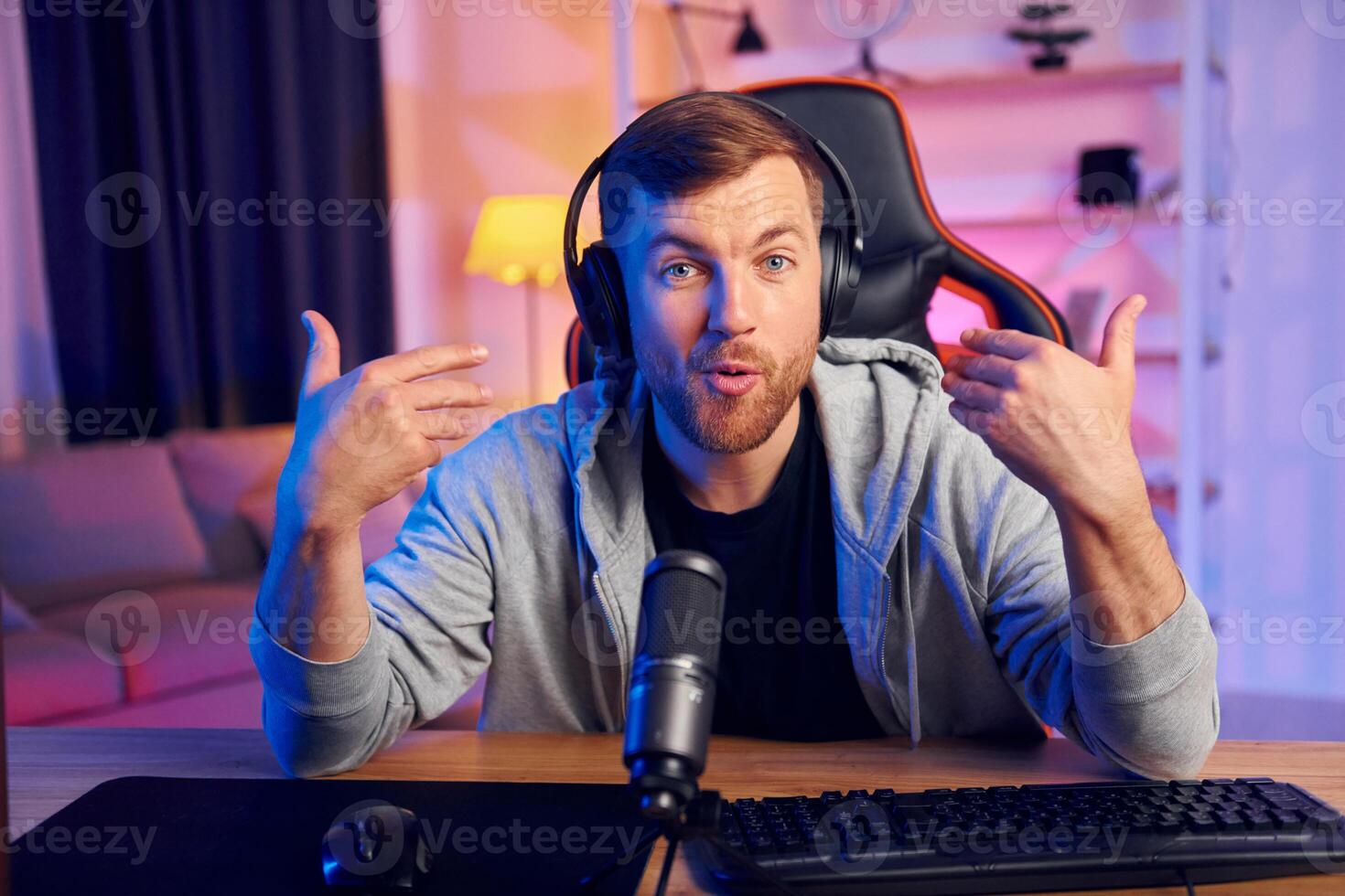 A man in a hoodie is sitting at a desk with headphones on photo