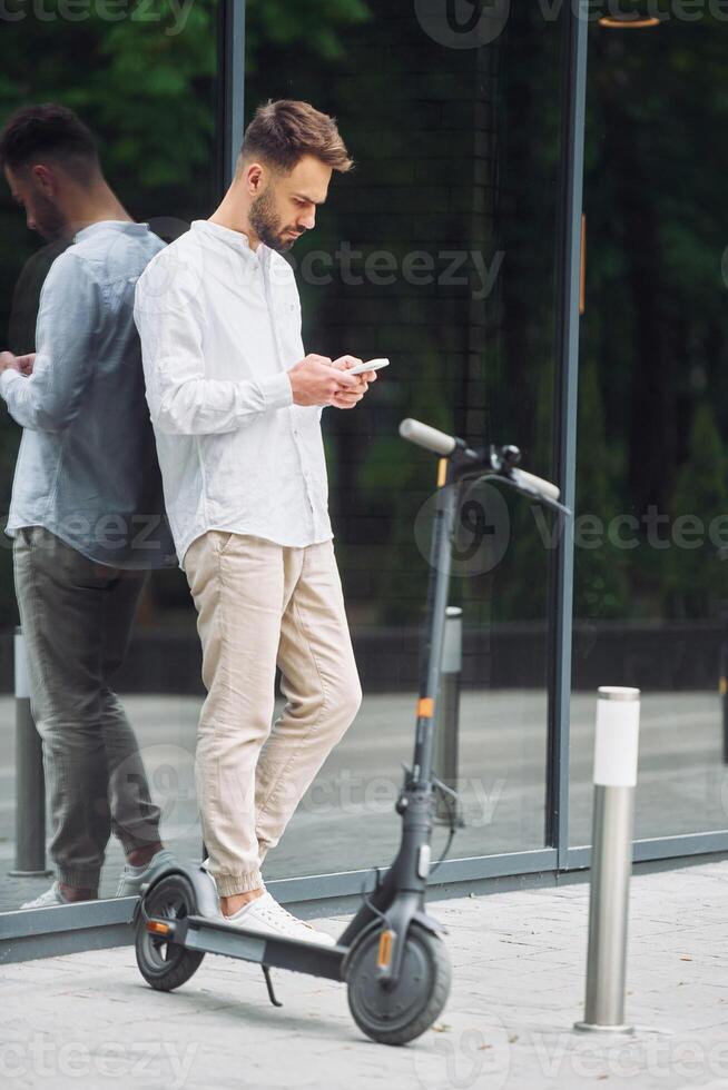 A man is standing on a scooter looking at his phone photo