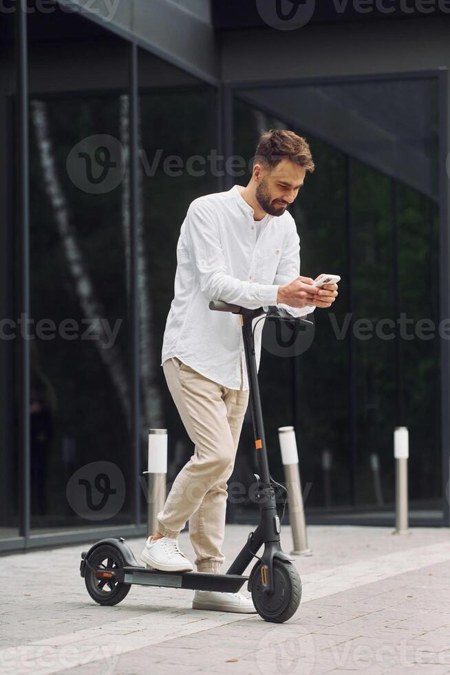 A man is standing on a scooter and looking at his phone photo