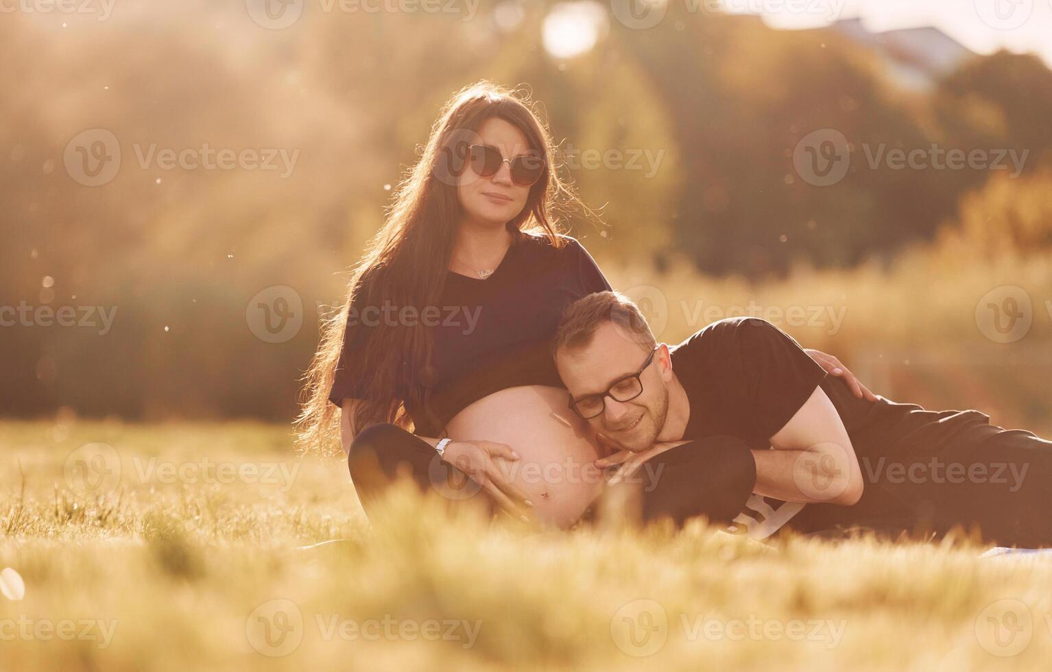 Man embracing his wife. Woman is pregnant. Lovely couple sitting on the field together photo