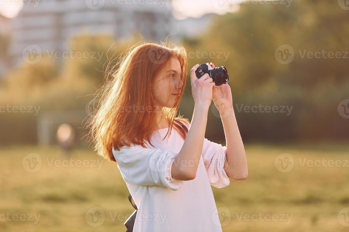 A Photographer is with camera. Young girl is on the field at sunny daytime having nice weekend photo