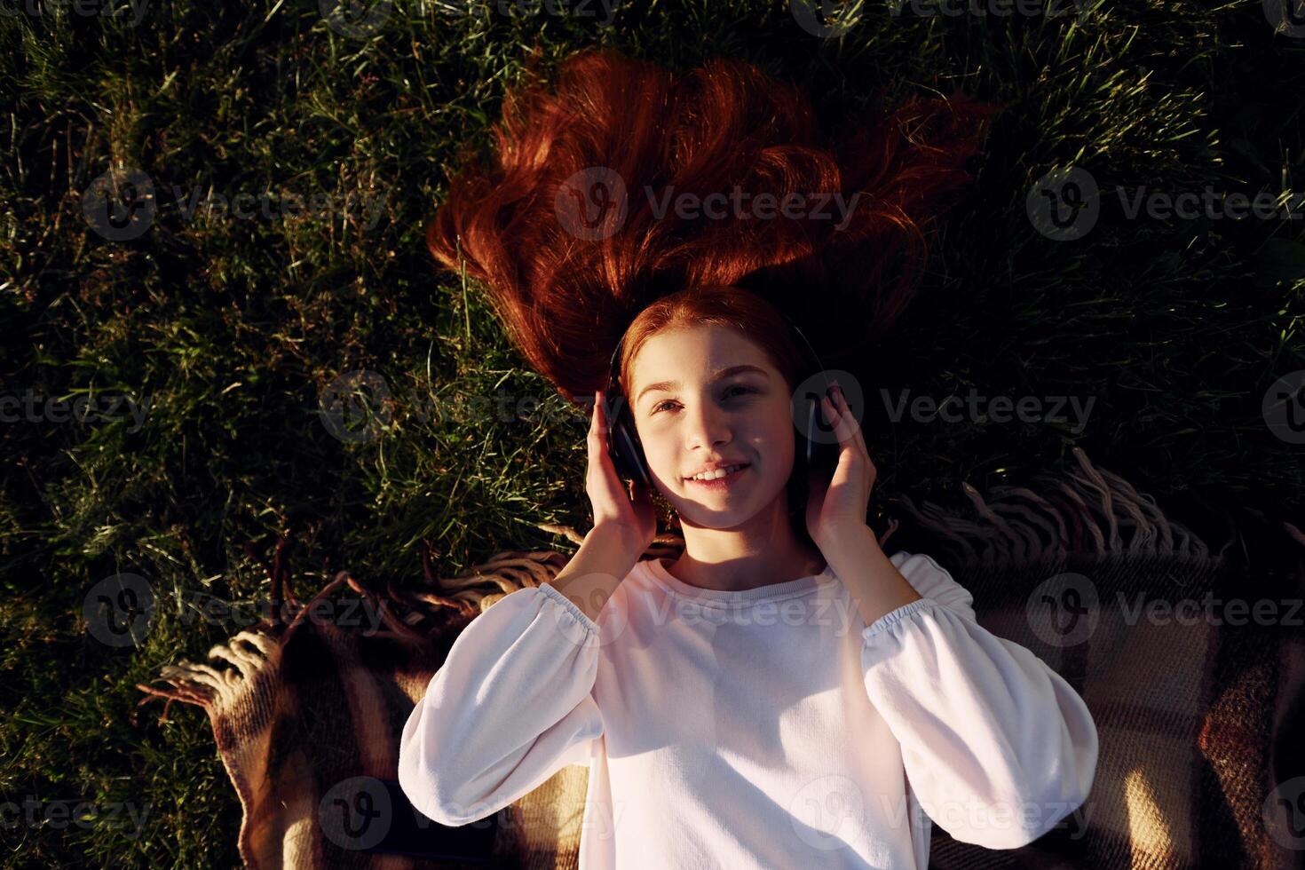 Top view. Listening to the music ,in headphones. Young girl is on the field at sunny daytime having nice weekend photo