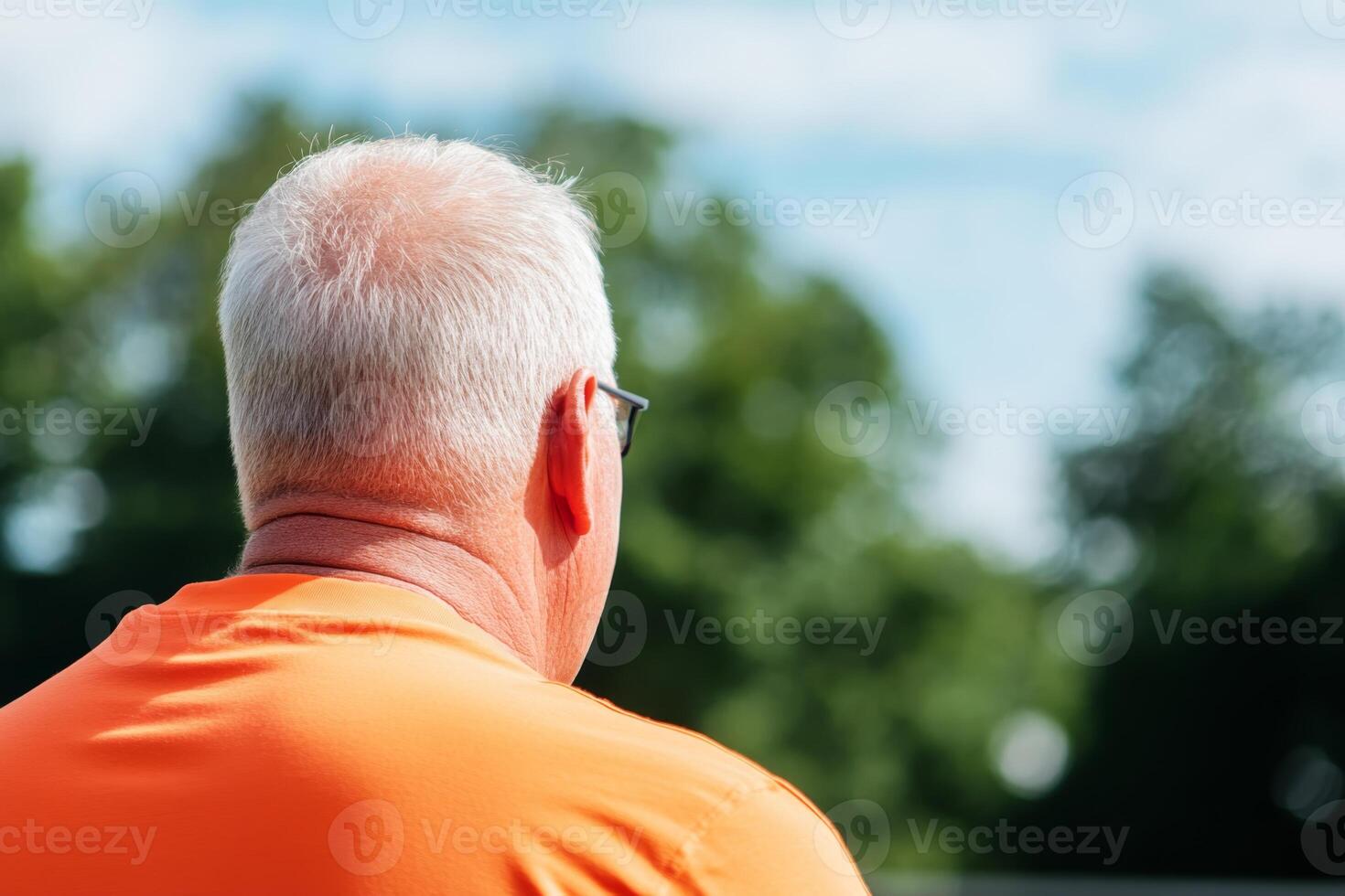 Man in orange shirt observes outdoor activity on a sunny day while surrounded by greenery photo