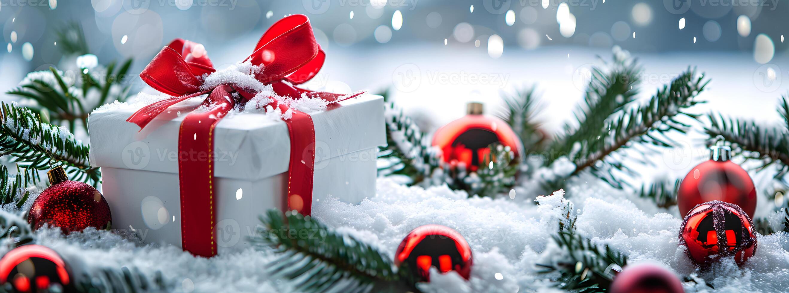 Beautifully wrapped gift box with red ribbon and bow, surrounded by pine branches and Christmas ornaments on soft, fresh snow photo