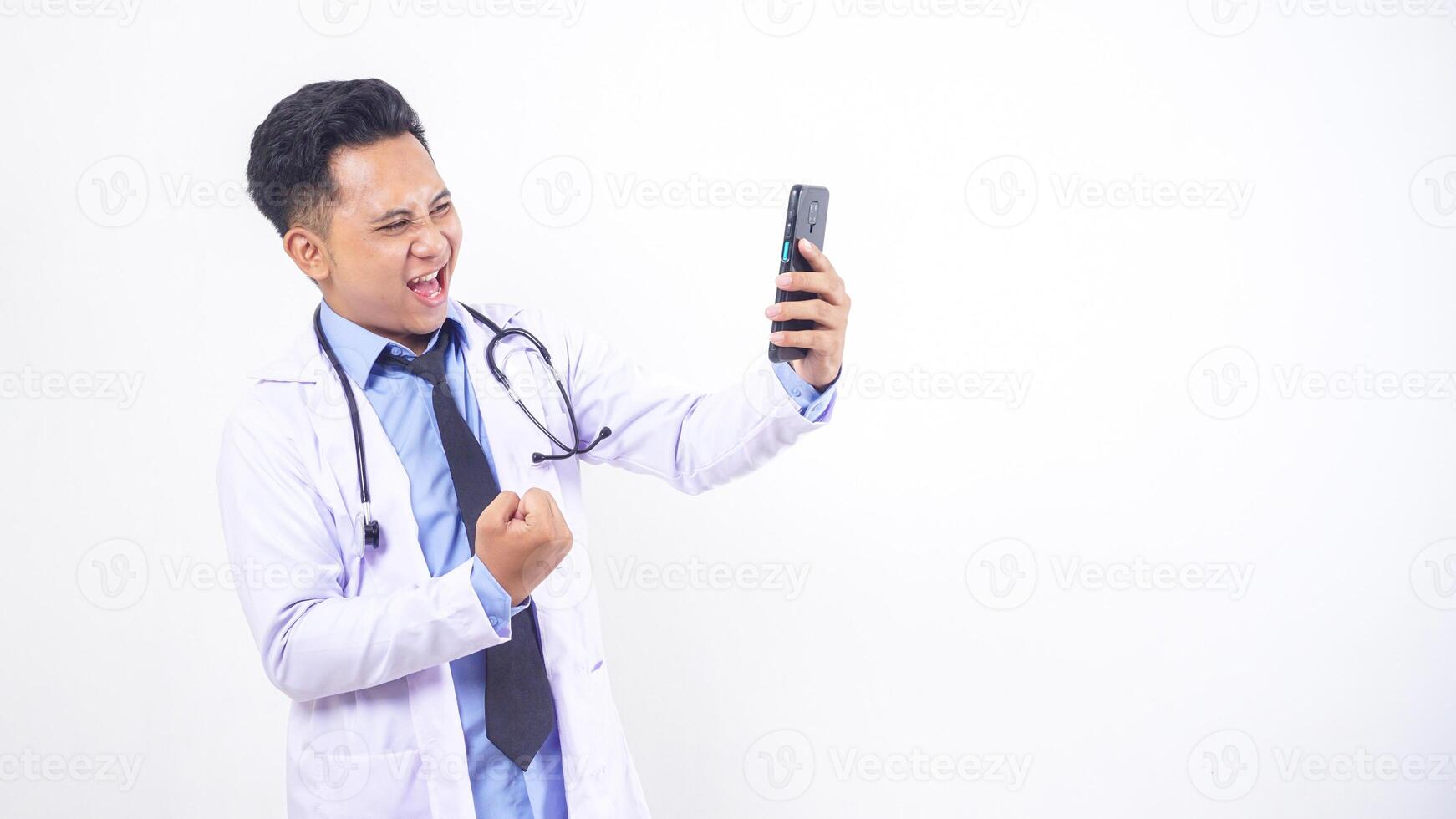 Excited Asian Male Doctor Holding Mobile Phone and Gesturing Yes Celebrating Successful on White Background photo