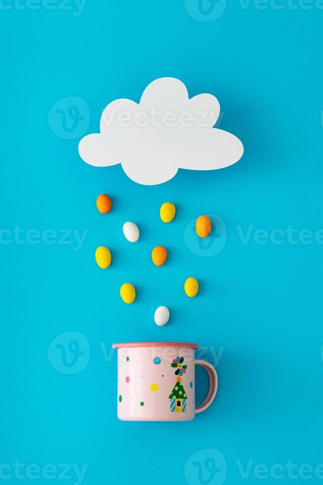 Creative composition made of cloud shape paper card note, colorful Easter eggs rain and pink enamel mug on blue background. Minimal Easter concept. Festive holiday layout. Easter aesthetic. Flat lay. photo