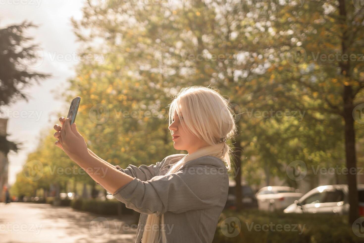 A woman is taking a picture of herself with her cell phone. She is wearing a gray jacket and scarf. The scene is set in a city with trees and cars in the background. photo
