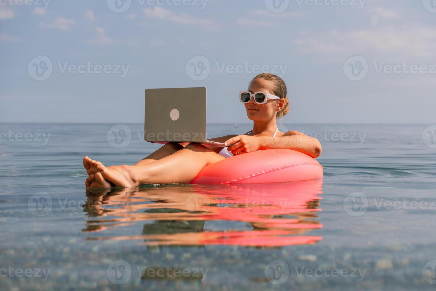 A woman is floating on a pink inflatable raft in the ocean while holding a laptop. Concept of relaxation and leisure, as the woman is enjoying her time in the water while working on her laptop. photo