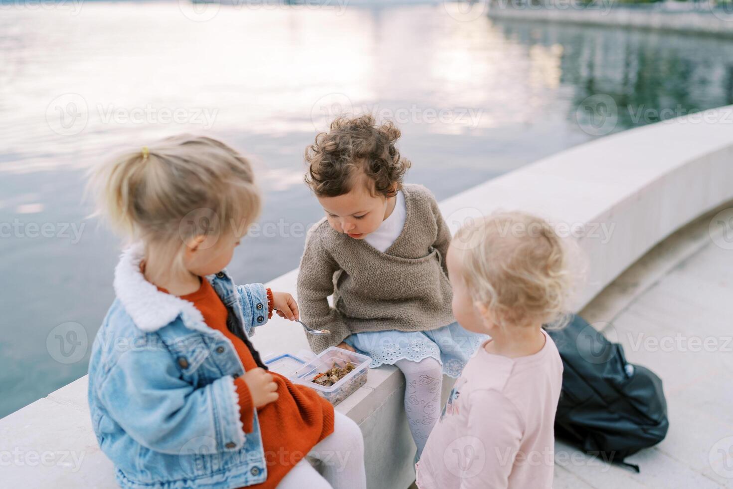 Little girl hands a spoon of porridge to her friend sitting with her on a fence by the sea photo