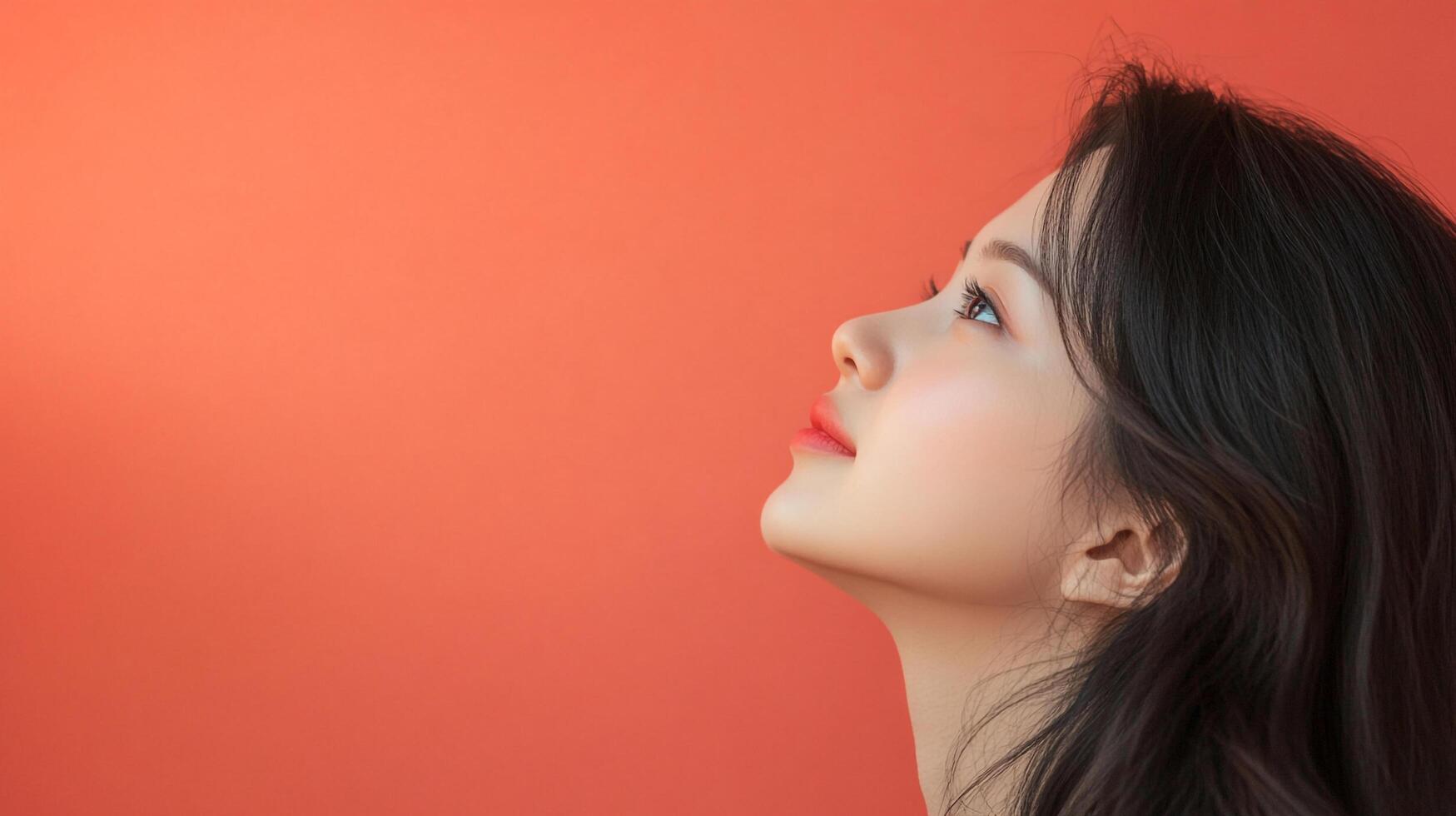 A young woman with soft, flowing dark hair looks upwards against a vibrant coral backdrop, showcasing beauty and serene contemplation. photo