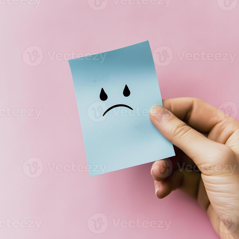 hand holding blue sticky note with sad face emoji against pink background conveys feelings of sadness and disappointment photo