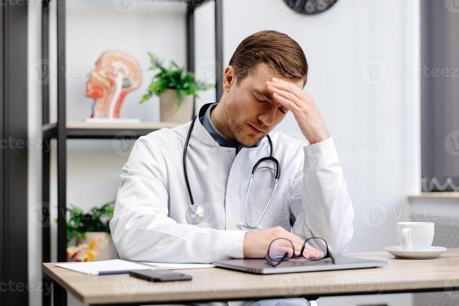 Doctor man wearing medical uniform sitting on his workplace tired holding his head feeling fatigue and headache. Stress and frustration concept. photo
