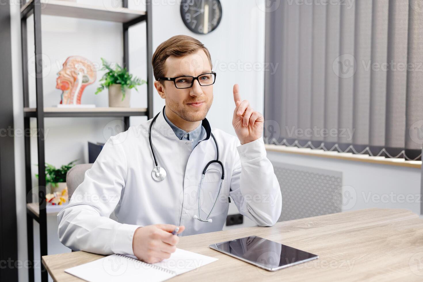 A young doctor in glasses and a white uniform sits at a desk and consults a patient online. The doctor asks for attention by raising his finger up photo