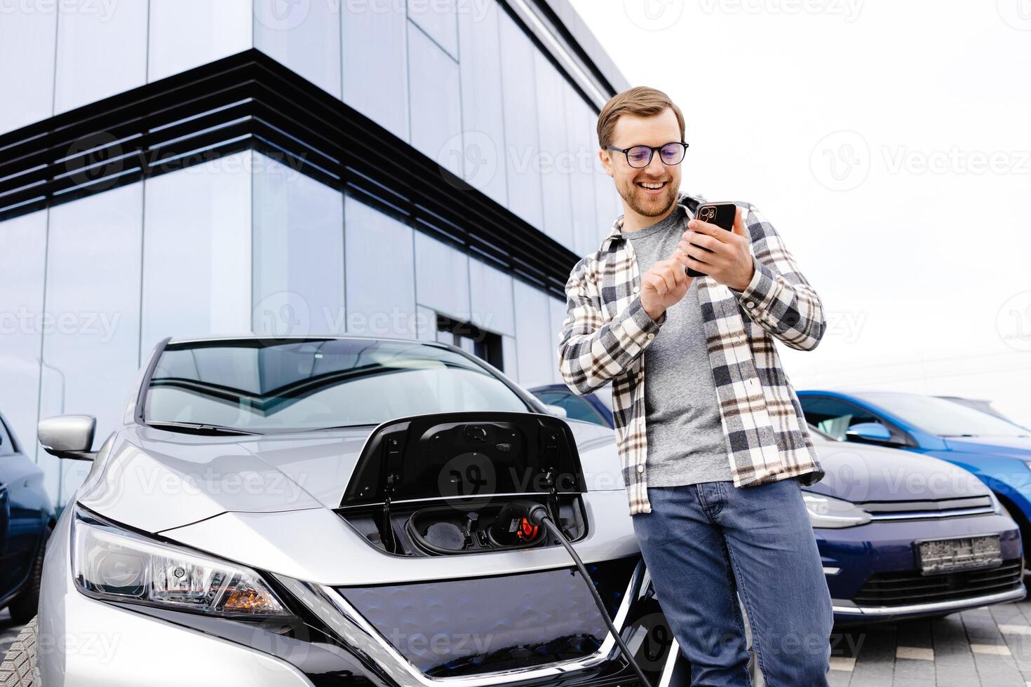 Young man uses a mobile phone while leaning on his electric car while the car is charging photo