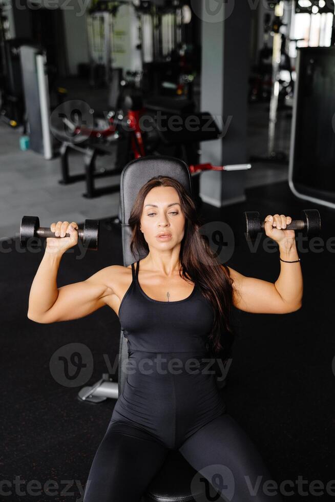 Young fit woman practicing dumbbell press while sitting on a bench. Slim female in sportswear exercises with weights in a gym. photo
