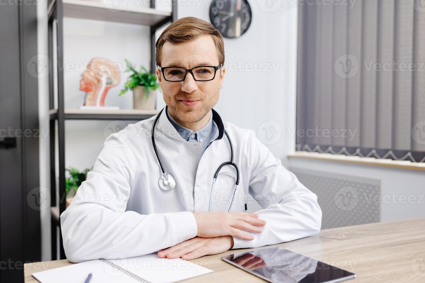Young doctor wearing glasses and white uniform with stethoscope, speaking and consulting patient online, looking at camera, making call, sitting at table in office photo