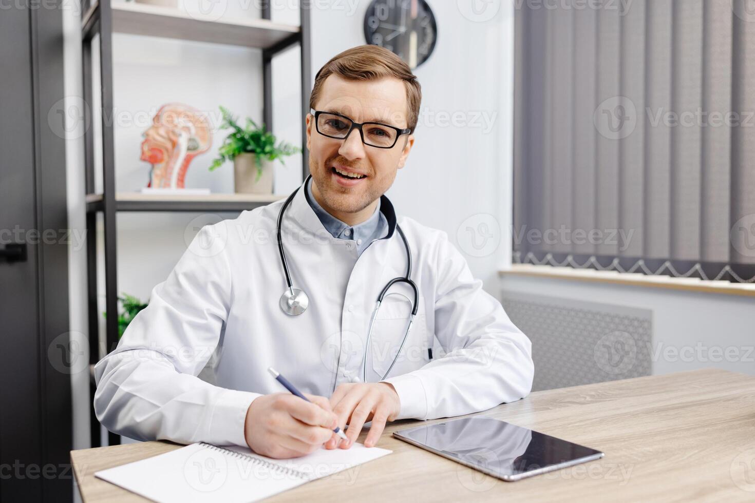 Doctor wearing glasses and white uniform with stethoscope, speaking and consulting patient online, looking at camera, making call, sitting at table in office. photo
