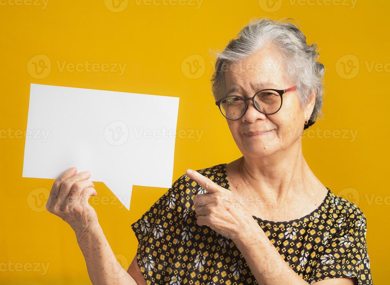 Senior woman holds and points to a blank speech bubble and looks at the camera with a smile photo