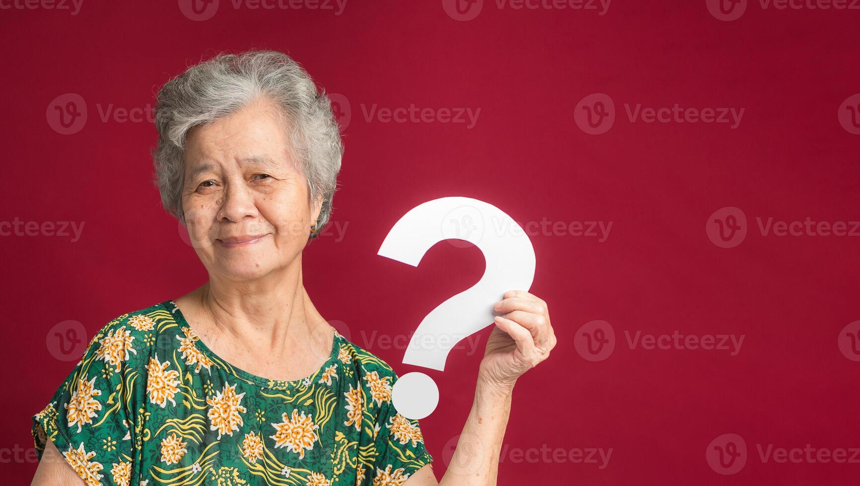 Senior woman holding a white question mark symbol with a smile while standing on a red background photo