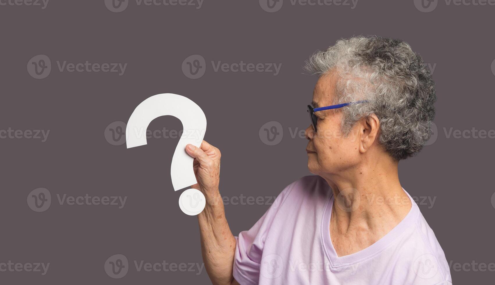 Senior woman holding a white question mark symbol with a smile while standing on a gray background photo
