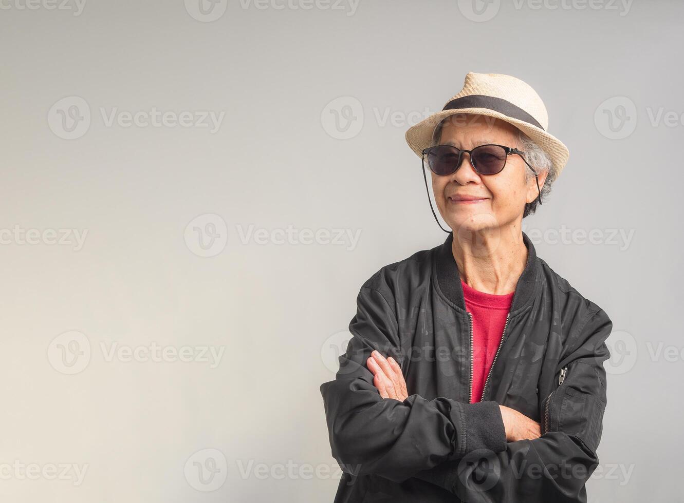 Beautiful elegant elderly woman in a black jacket wearing sunglasses and a hat with a smile while standing with arms crossed on a gray background photo