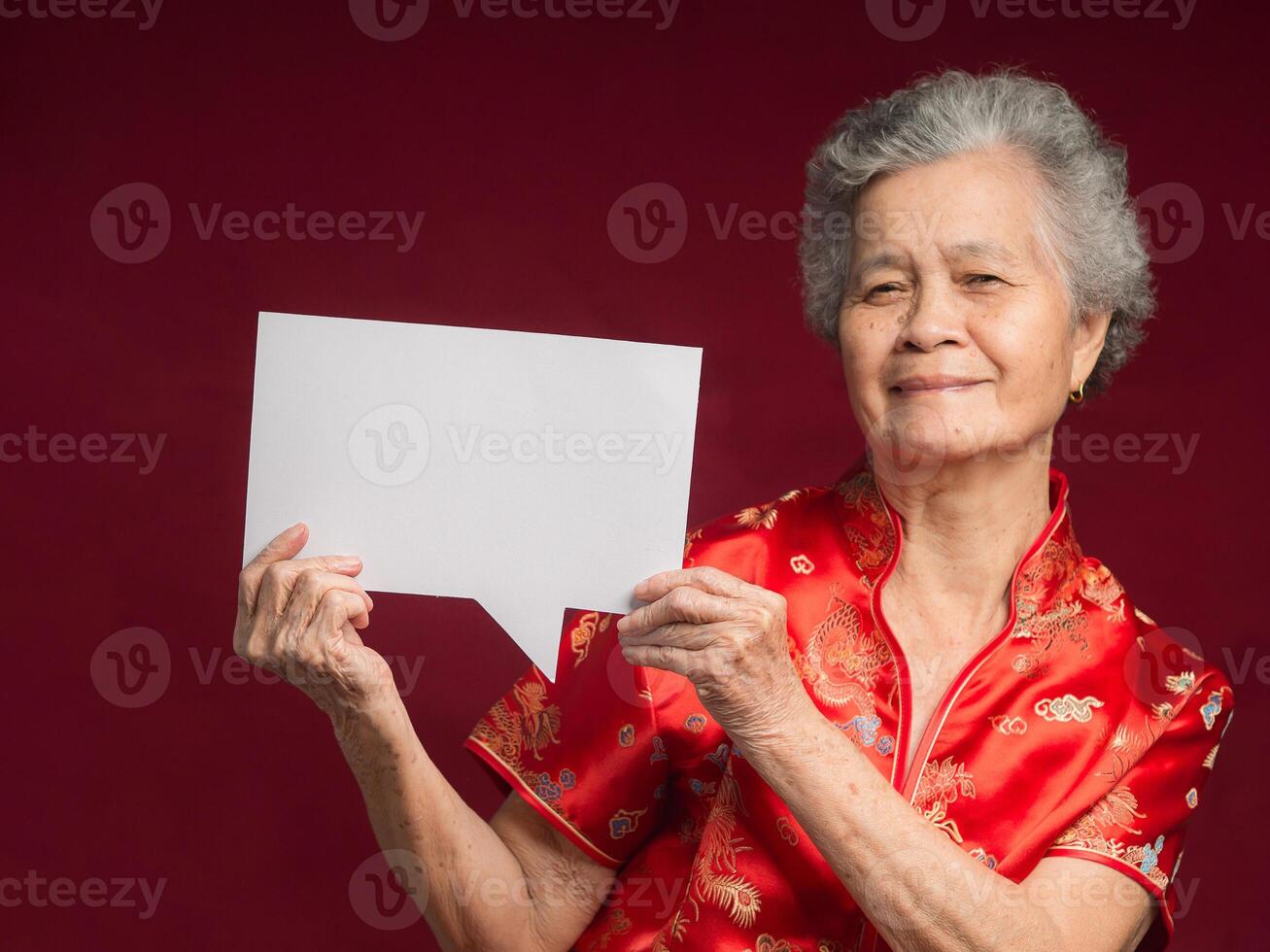 Portrait of a senior woman wearing a traditional cheongsam qipao holding a blank speech bubble while standing on a red background photo