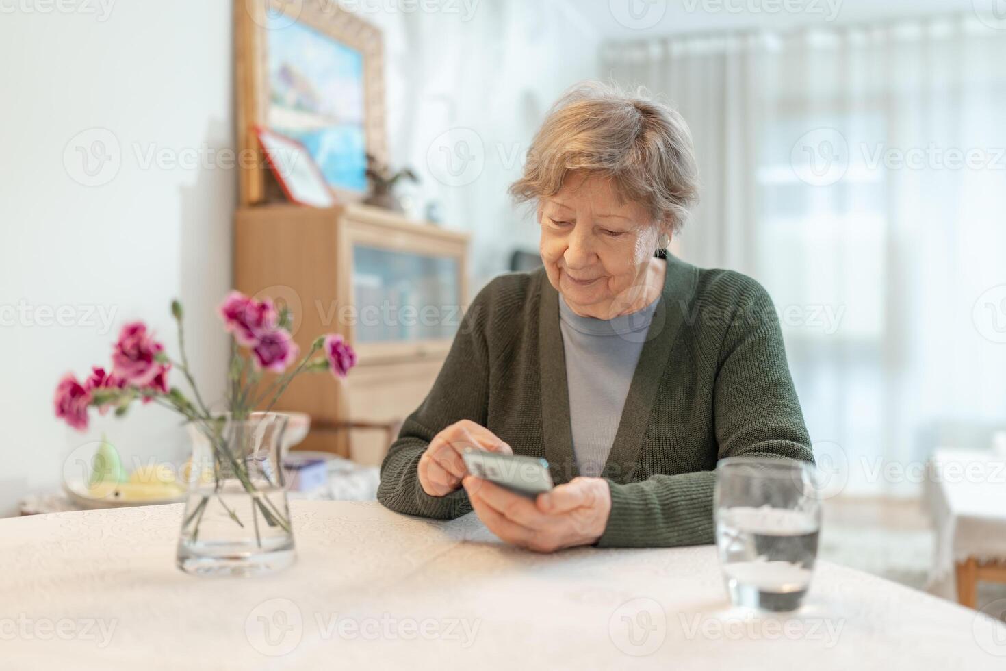 A senior woman uses a smartphone while sitting at a table in a cozy home environment. Concept of technology, digital literacy, online communication, independent learning, and active aging. photo