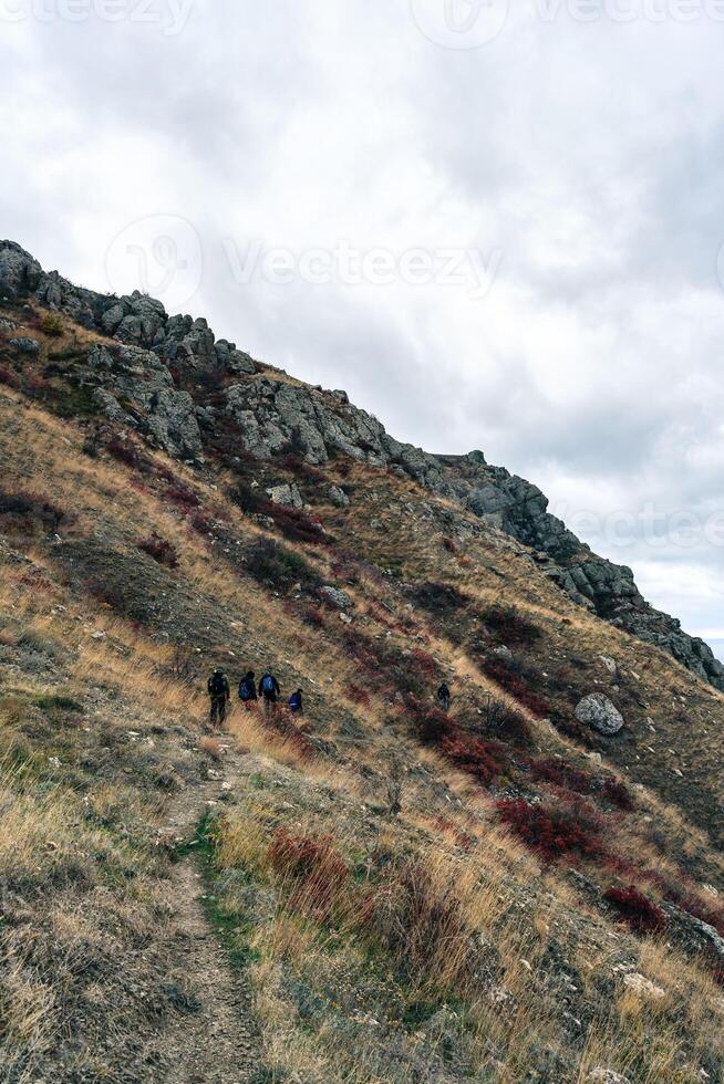 Group of hikers trekking along a rocky hillside under a cloudy sky in a mountainous region during autumn photo