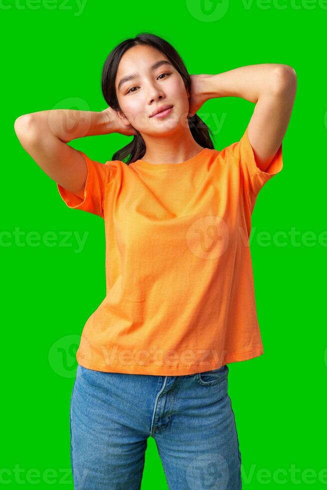 Young woman poses confidently against a bright green background while wearing a casual orange t-shirt and denim jeans photo