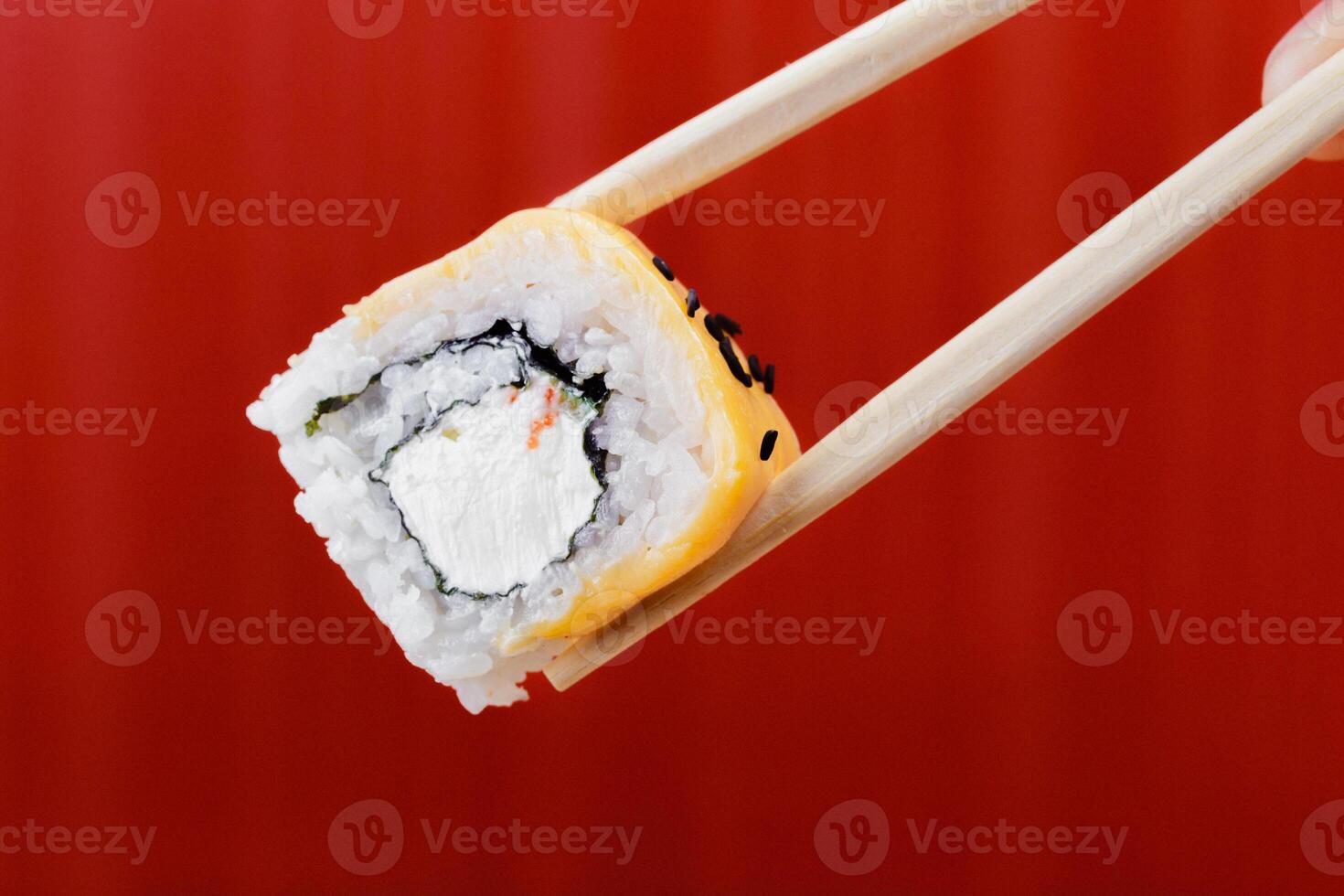 Sushi roll with cream cheese and crab held by chopsticks in front of a red background photo