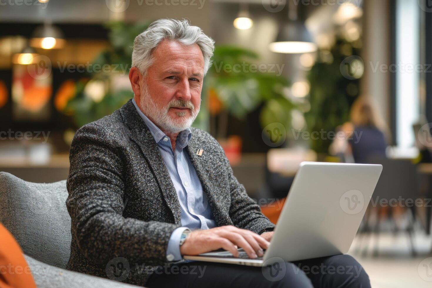 Man sits typing on silver laptop. Older man works on computer. Business person looks at camera. Working indoors in casual clothes. Grey hair. photo