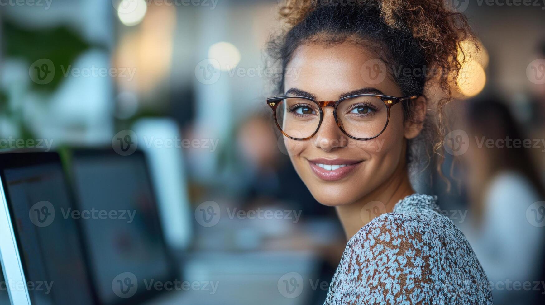 Office worker with glasses. Young woman smiles in workspace. Computer screen and other people in background. Good for professional or business theme. photo