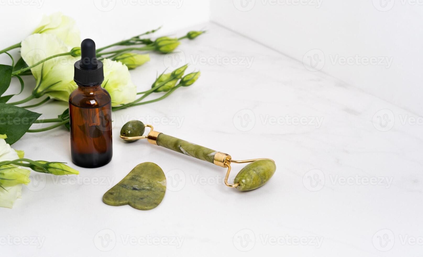 Jade roller for face massage. Green gua sha on the marble white background with flat lay. Anti age, lifting and toning treatment, acupressure. Eustoma flowers. Serum brown bottle pipette photo