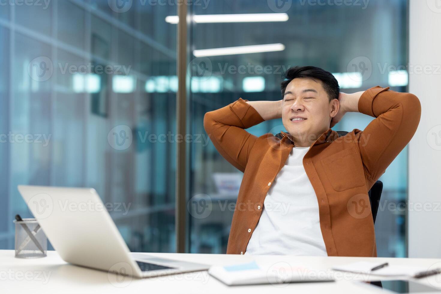 Man resting and relaxing at workplace inside office. Satisfied Asian man put his hands behind his head with closed eyes smiling, successful achievement results, well done. photo