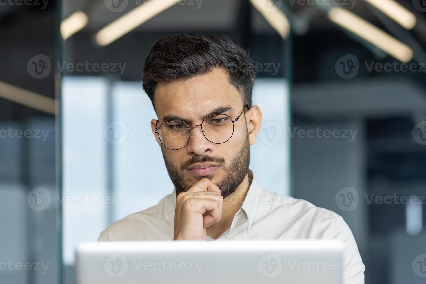Man wearing glasses concentrating on computer screen in office setting. Serious expression suggests deep thought or analysis. Modern work environment. Keywords. office, focus, professional. photo