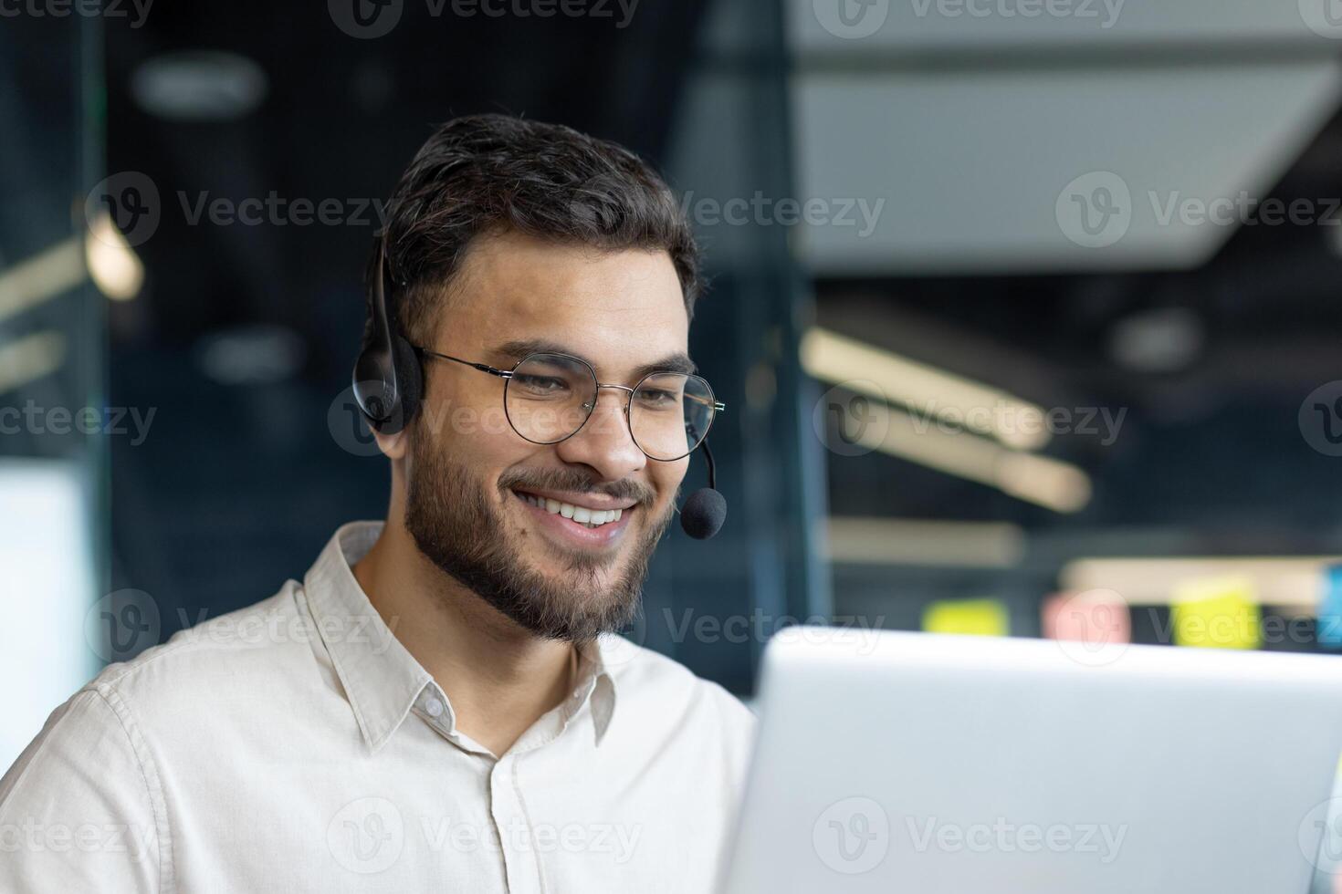A man wearing glasses and a headset smiles while working on his laptop, embodying a cheerful and dedicated office professional in a modern work setting. photo