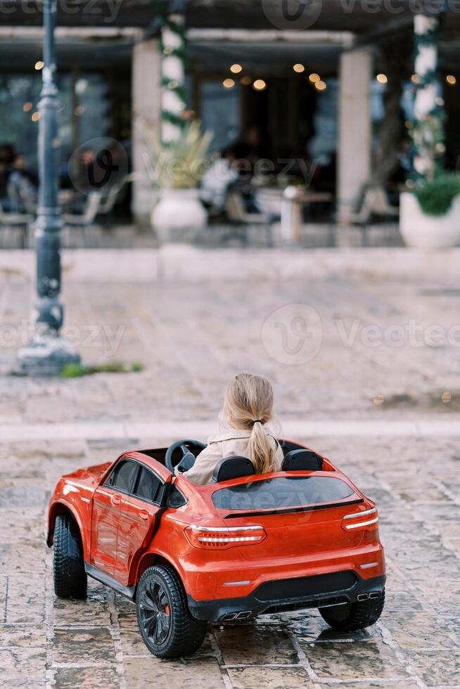 Little girl drives a red toy car down the street. Back view photo