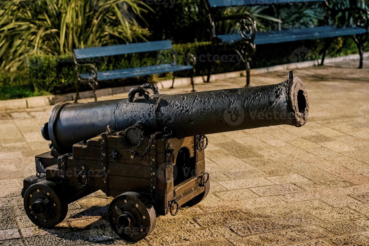 Old bronze cannon on a wooden cart with wheels stands on the paving stones near the bench photo