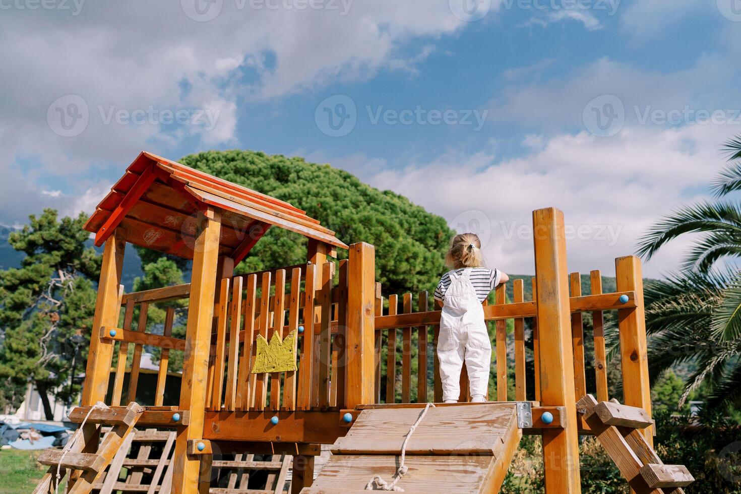 Little girl stands on a wooden slide on a playground, leaning on a fence. Back view photo