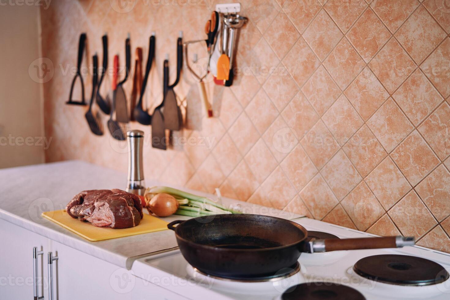 Frying pan stands on the stove in the kitchen next to raw meat and vegetables on the table photo