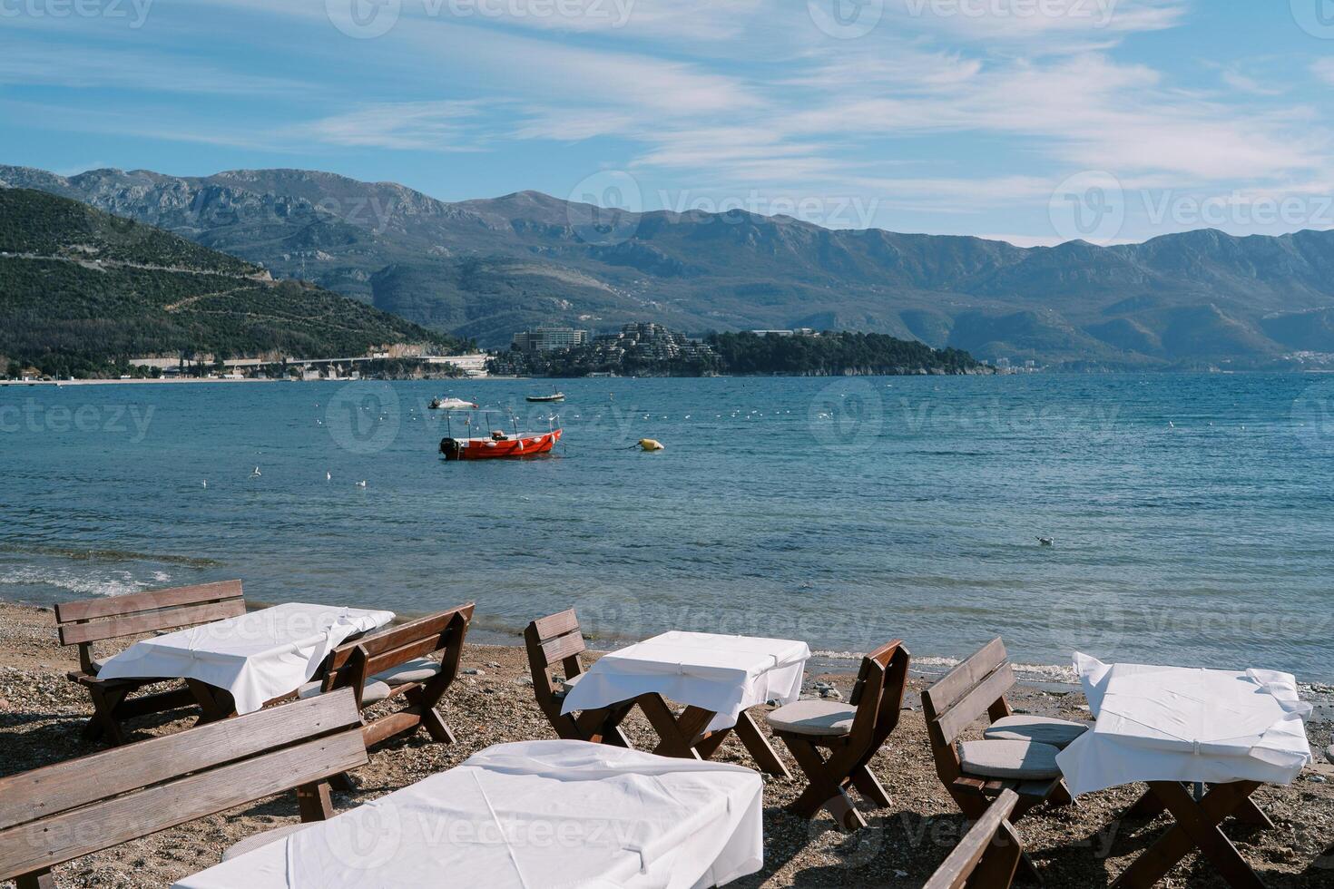 Wooden tables with benches stand on a sandy beach near the sea with a moored red boat photo