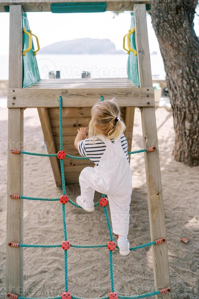 Little girl climbs a rope ladder onto a slide on a beach playground. Back view photo