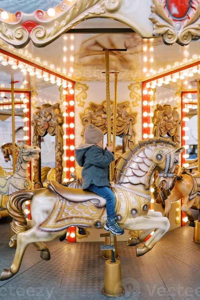 Little girl rides a horse on a colorful carousel holding the handle. Side view photo