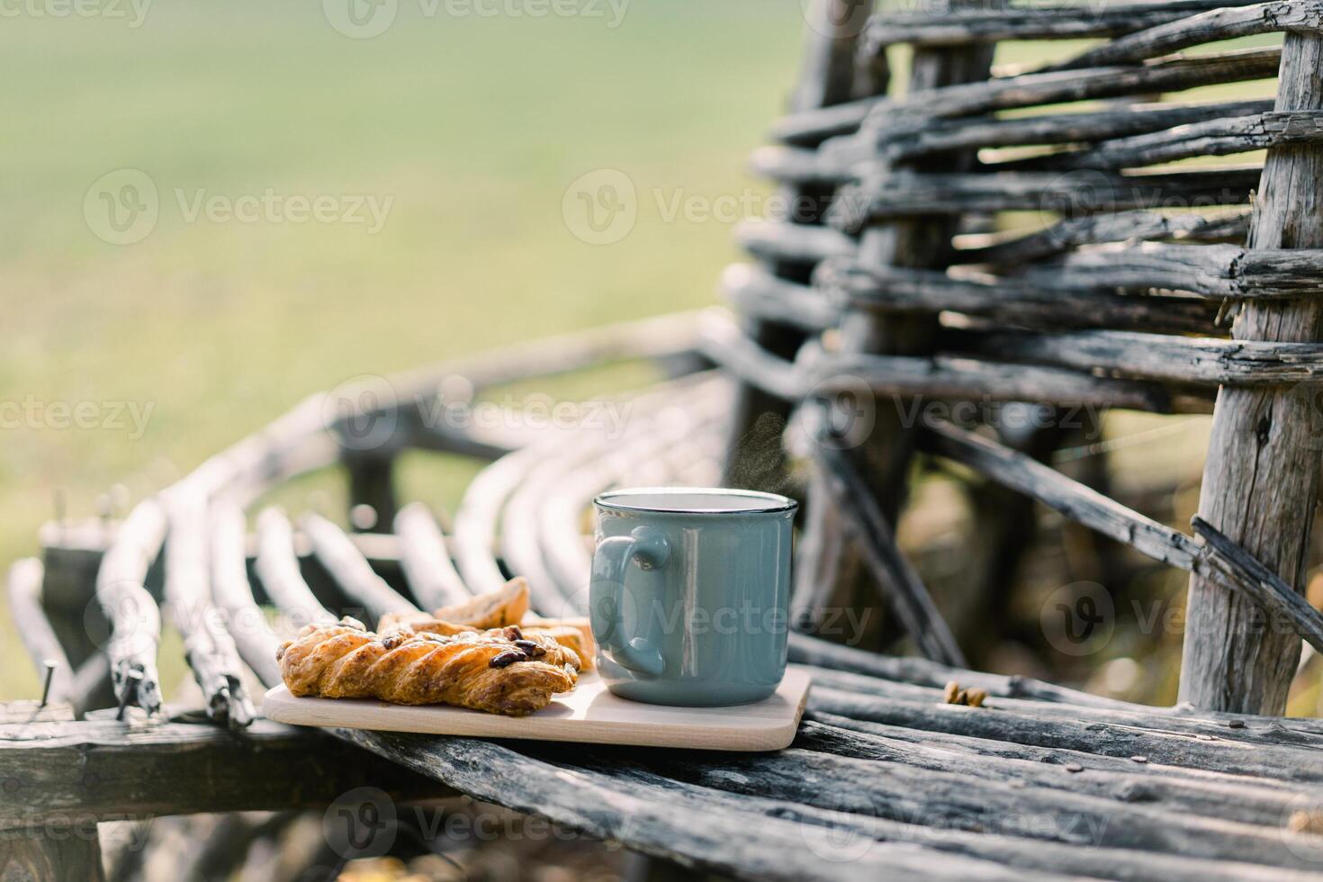 Mug of coffee next to a bun stands on a wooden plank on a park bench photo