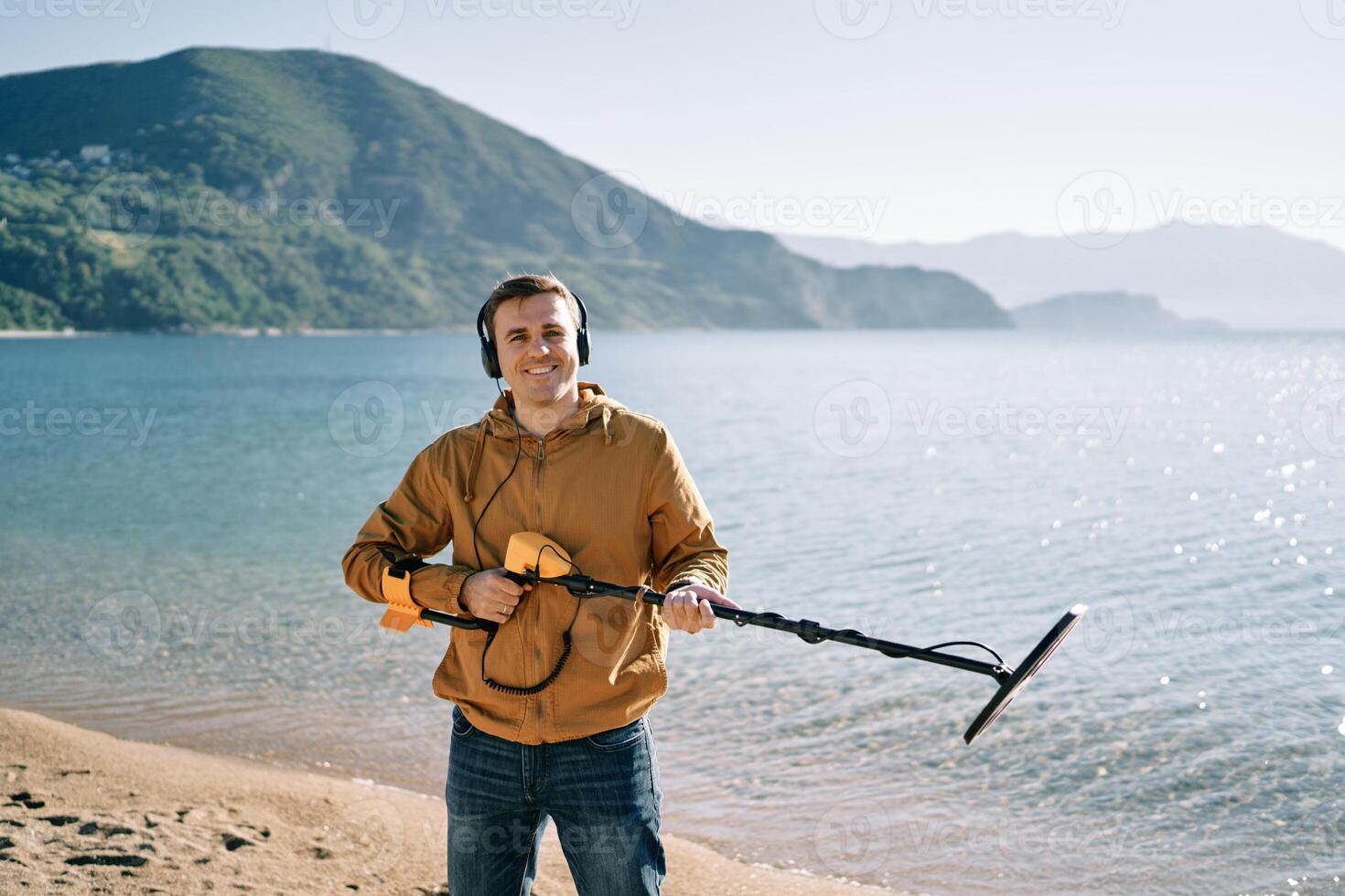 Smiling man with a metal detector at the ready stands on the seashore photo
