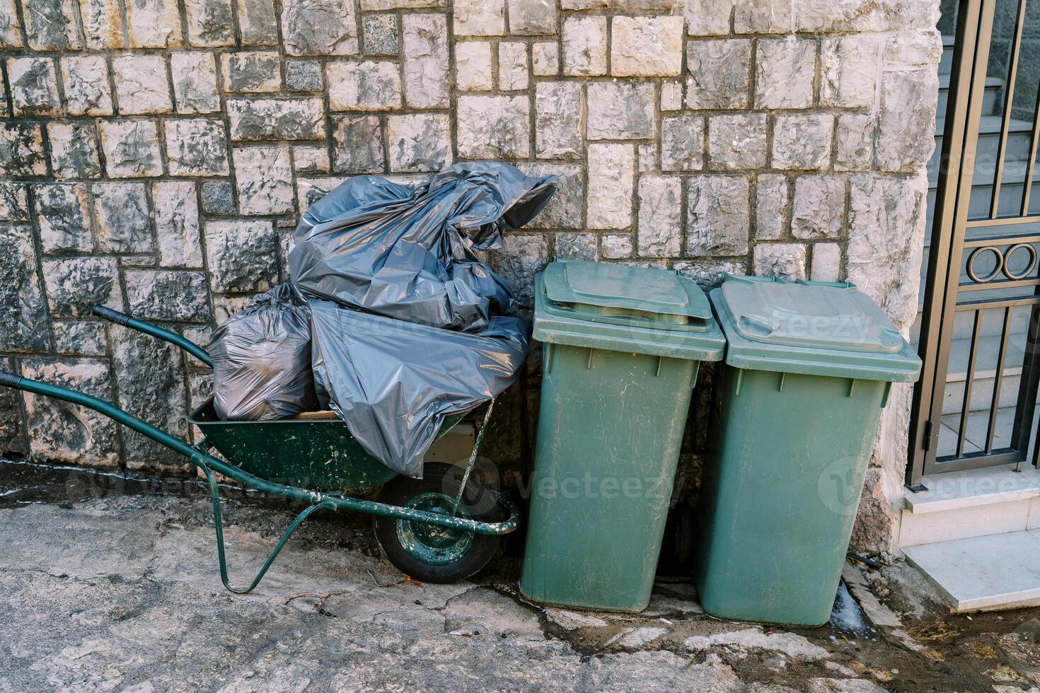 Cart with bags of garbage stands near plastic garbage containers near a stone fence photo