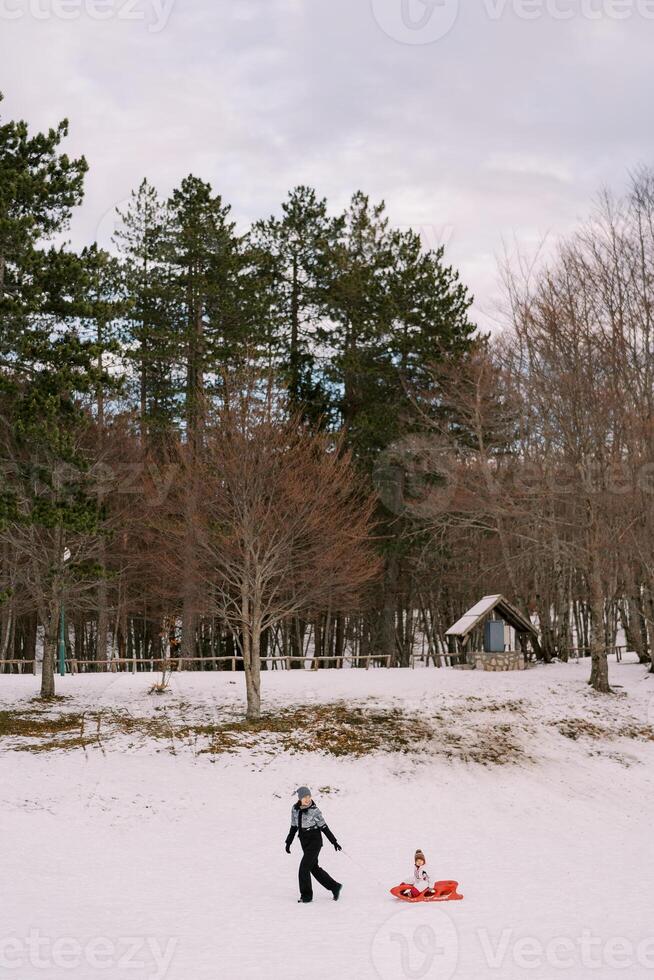 Mom is carrying a small child in a sled past a wooded hill with a stone shed. Side view photo
