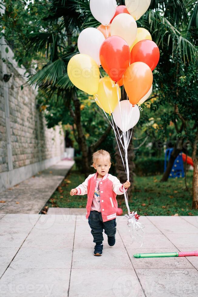Little girl with a bunch of colorful balloons walks through a green garden photo